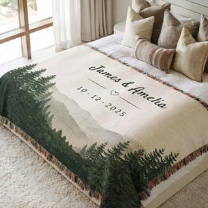 Puede incluir: Una manta tejida de color crema con un diseño de bosque y montaña. La manta presenta los nombres "James & Amelia" y la fecha "10-12-2025". La manta tiene un borde con flecos y se muestra en una cama con almohadas.