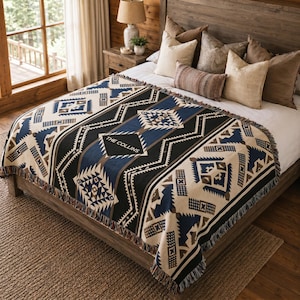 May include: A woven blanket with a geometric pattern in black, blue, and beige, draped over a wooden bed frame. The blanket features a fringed edge and the text "THE COLLINS" in a diamond shape.