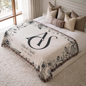 Puede incluir: Manta crema con borde floral negro y flecos. La manta muestra las iniciales "AS", los nombres "Allen & Sophia" y la fecha "05-24-2024". La manta está sobre una cama con almohadas.