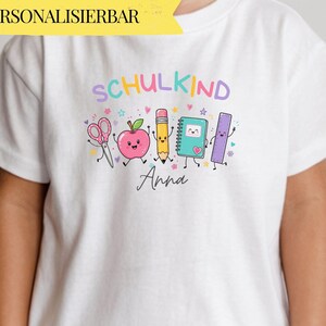 Puede incluir: Una camiseta blanca de manga corta con un gráfico colorido de útiles escolares de dibujos animados, incluyendo tijeras, una manzana, un lápiz, un cuaderno y una regla. El texto dice SCHULKIND y el nombre Anna en letra cursiva debajo.