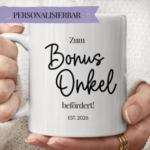 Peut inclure: Un mug en céramique blanche avec le texte noir Zum Bonus Onkel befördert! EST. 2026. Une bannière violette en haut contient le mot PERSONALISIERBAR. Le mug présente une anse incurvée et une finition lisse et brillante avec une écriture sombre.