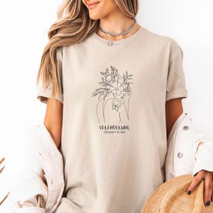 Peut inclure: T-shirt beige avec un dessin au trait d'une femme ornée de fleurs et de papillons. Le texte "VERÄNDERUNG BEGINNT IN MIR" est imprimé en dessous. Le t-shirt a des manches courtes et une coupe ample.