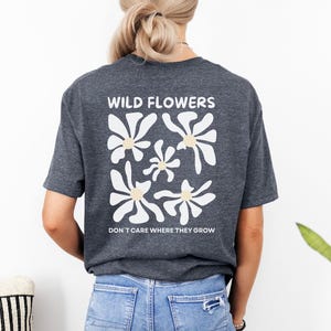 Peut inclure: T-shirt gris foncé avec un motif floral au dos. Le motif présente des fleurs blanches avec des centres jaunes et le texte "WILD FLOWERS" au-dessus et "DON'T CARE WHERE THEY GROW" en dessous.