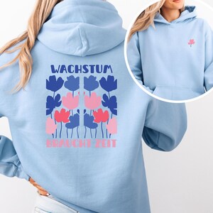 Peut inclure: Sweat à capuche bleu clair avec un motif floral au dos. Le motif présente des fleurs bleues et roses avec le texte allemand "WACHSTUM BRAUCHT ZEIT". Une petite fleur rose est sur le devant. Le sweat à capuche a une capuche et une poche avant.