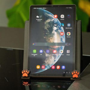 Könnte beinhalten: Ein schwarzes Tablet mit einem farbenfrohen Erdbild, das in einem schwarzen Ständer mit orangefarbenen Pfotenabdruck-Akzenten steht. Der Tablet-Bildschirm zeigt App-Symbole und die Google-Suchleiste. Der Ständer steht auf einer dunklen Oberfläche.