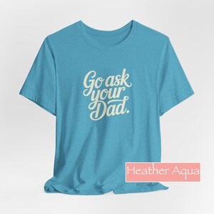 May include: A turquoise t-shirt featuring the text "Go ask your Dad" in a cream-colored, cursive font. The shirt is a crew neck style. The label at the bottom right says "Heather Aqua".