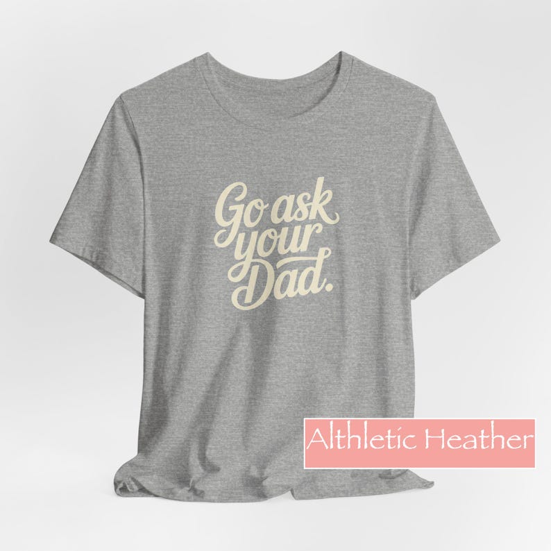 May include: A grey t-shirt featuring the text "Go ask your Dad" in a cursive font. The shirt has short sleeves and a crew neck. The text "Althletic Heather" is in a pink rectangle at the bottom.