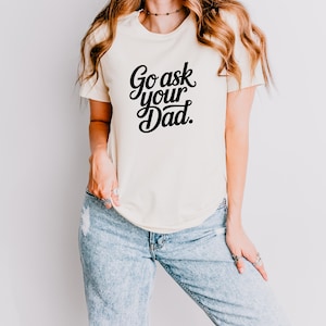 May include: A cream-coloured t-shirt featuring the text "Go ask your Dad" in black script. The shirt is worn with light blue ripped jeans. The style is casual and relaxed.