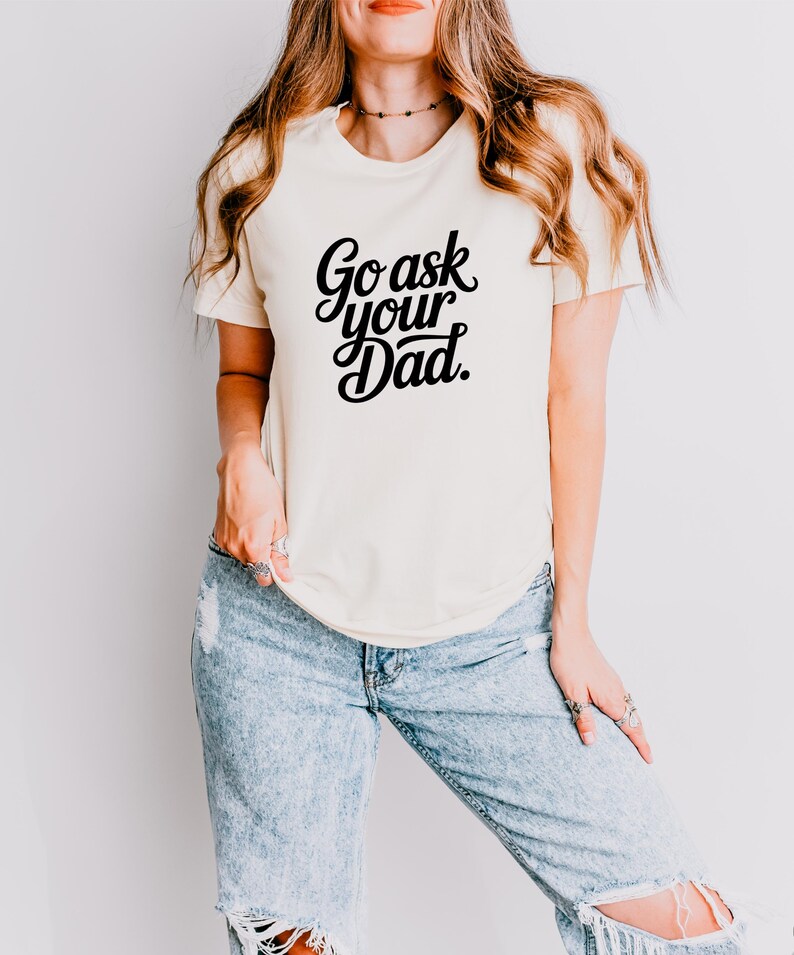 May include: A cream-coloured t-shirt featuring the text "Go ask your Dad" in black script. The shirt is worn with light blue ripped jeans. The style is casual and relaxed.