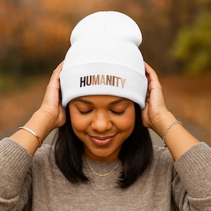 Może przedstawiać: Biała czapka z napisem "HUMANITY" w brązowych literach. Czapka jest trzymana przez osobę. Tło jest rozmyte z jesiennymi kolorami.