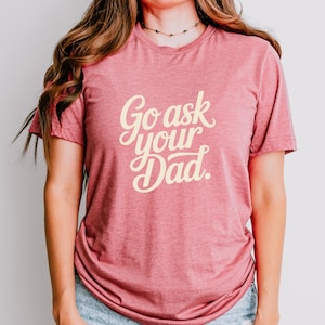 May include: A red t-shirt with the text "Go ask your Dad" in a cursive font. The shirt is made of a soft material and is being worn by a person. The person is wearing blue jeans.