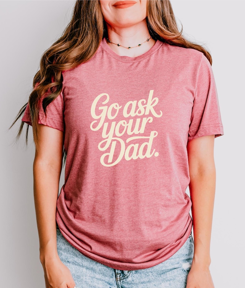May include: A red t-shirt with the text "Go ask your Dad" in a cursive font. The shirt is made of a soft material and is being worn by a person. The person is wearing blue jeans.