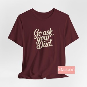 May include: Maroon t-shirt with the text "Go ask your Dad" in a cursive font. The shirt is a solid maroon colour and has short sleeves. The word "Maroon" is in a pink box in the bottom right corner.