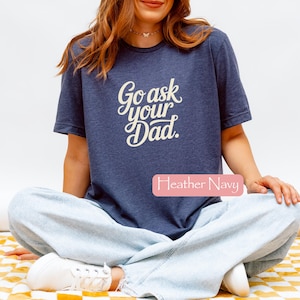 May include: A navy blue t-shirt with the text "Go ask your Dad" in a cursive font. The shirt is worn by a person sitting cross-legged. The words "Heather Navy" are in a pink rectangle.