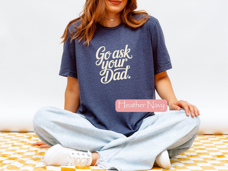 May include: A navy blue t-shirt with the text "Go ask your Dad" in a cursive font. The shirt is worn by a person sitting cross-legged. The words "Heather Navy" are in a pink rectangle.