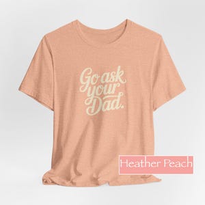 May include: A heather peach-coloured t-shirt with the text "Go ask your Dad" in a cursive font. The shirt is a short-sleeved crew neck style. The colour is described as "Heather Peach" in a rectangular box at the bottom.