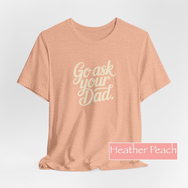 May include: A heather peach-coloured t-shirt with the text "Go ask your Dad" in a cursive font. The shirt is a short-sleeved crew neck style. The colour is described as "Heather Peach" in a rectangular box at the bottom.