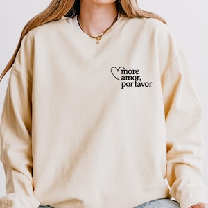 May include: A cream-coloured sweatshirt featuring the phrase "more amor, por favor" in black script and a heart graphic. The long-sleeved top is a neutral colour and has a relaxed fit. The text is on the upper left chest area.