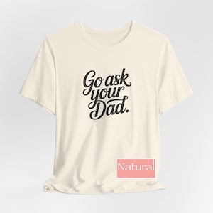 May include: A cream-coloured t-shirt featuring the black script text "Go ask your Dad". The shirt has a crew neck and short sleeves. The word "Natural" is displayed in a pink rectangle at the bottom.