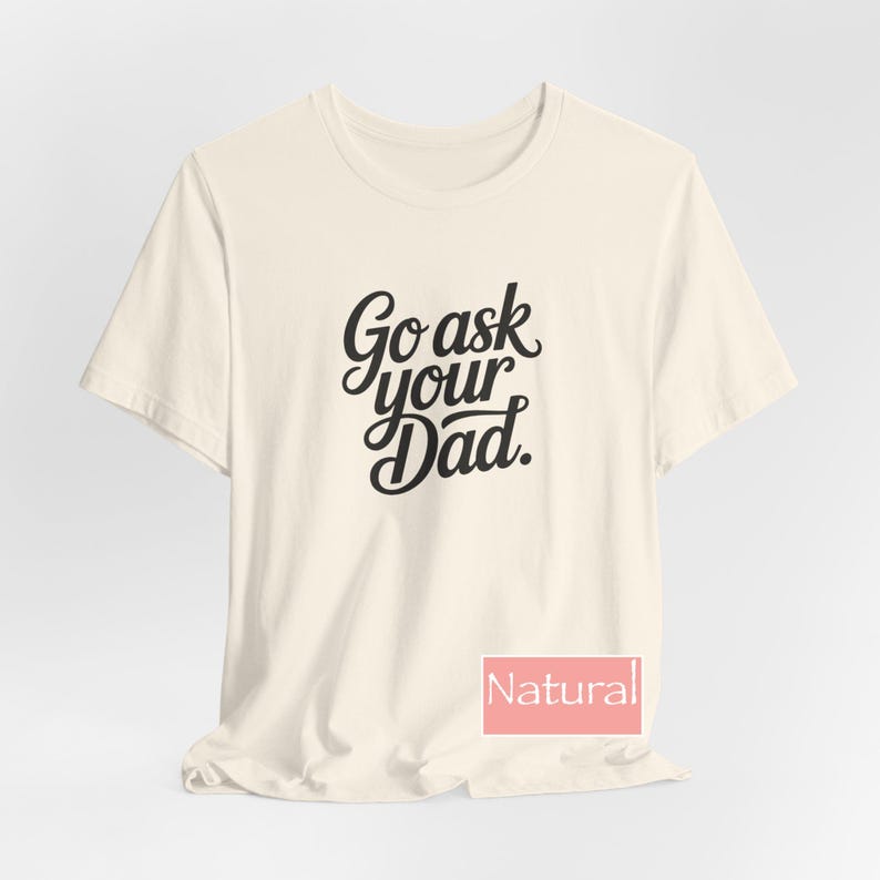 May include: A cream-coloured t-shirt featuring the black script text "Go ask your Dad". The shirt has a crew neck and short sleeves. The word "Natural" is displayed in a pink rectangle at the bottom.