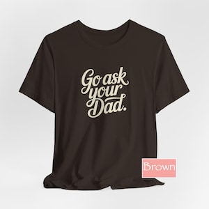 May include: A brown t-shirt featuring the text "Go ask your Dad" in an off-white, cursive font. The shirt has short sleeves and a solid colour. The word "Brown" is in a pink box in the bottom right corner.