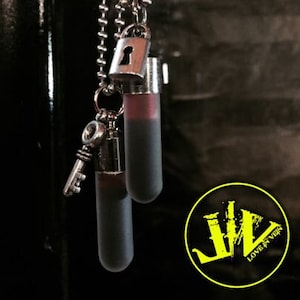 May include: A silver-toned necklace featuring a small padlock charm, a key charm, and two cylindrical vials. The vials contain a dark red liquid. The necklace hangs against a dark background. A yellow logo with the text "LOVE IN VEIN" is in the bottom right corner.