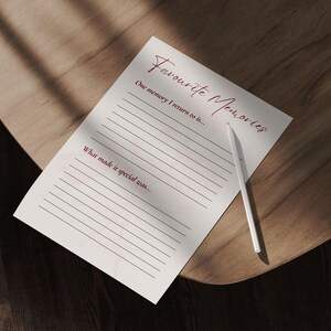 May include: A white paper with the words "Favourite Memories" in burgundy script, with lined spaces for writing. A white pencil rests on the paper, which is on a wooden surface. The paper prompts the user to write about a favorite memory.