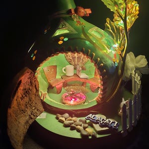 Puede incluir: Una casa en miniatura iluminada y caprichosa con un exterior verde, que presenta un interior acogedor con una mesa, sillas y una chimenea brillante. También se ve una gran flor iridiscente y la palabra "ALACATI".