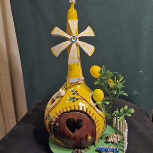 Puede incluir: Una caprichosa pieza de arte de calabaza amarilla pintada a mano. Presenta una puerta en forma de corazón, un molino de viento y un pequeño jardín con limones y una valla blanca. La calabaza está decorada con un pájaro y otros detalles.