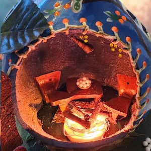 Puede incluir: Un diorama artesanal y caprichoso que presenta una escena interior en miniatura. La estructura azul y marrón tiene un interior acogedor con muebles diminutos, una chimenea y una mesa. El exterior está decorado con detalles florales.