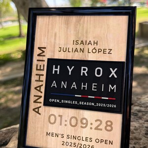 Puede incluir: Una placa rectangular en un marco negro con un fondo de veta de madera clara. El texto incluye ISAIAH JULIAN LÓPEZ, ANAHEIM, HYROX ANAHEIM, OPEN_SINGLES_SEASON_2025/2026, 01:09:28 y MEN'S SINGLES OPEN 2025/2026 con una línea roja, blanca y azul.