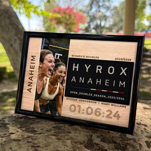 Puede incluir: Una placa rectangular enmarcada con una foto de dos atletas, el texto "ANAHEIM", "WOMEN'S DOUBLES", "2025/2026", "HYROX ANAHEIM", "OPEN_DOUBLES_SEASON_2025/2026", "BROOKE HENDERSON | MARY WILLIAMS" y la hora "01:06:24" impresos sobre madera.