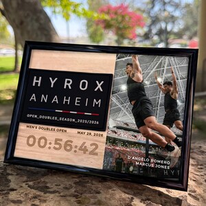 Puede incluir: Una placa rectangular enmarcada con fondo de veta de madera con el texto HYROX ANAHEIM, OPEN_DOUBLES_SEASON_2025/2026, MEN'S DOUBLES OPEN, MAY 29, 2026 y 00:56:42. Incluye una foto de dos atletas saltando y D'ANGELO ROMERO MARCUS JONES.