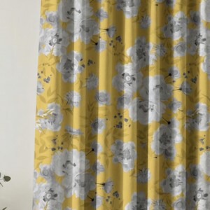 Puede incluir: Panel de cortina amarillo mostaza con un gran estampado floral en tonos grises y blancos. La cortina presenta un diseño repetido de flores y hojas estilizadas, creando un aspecto suave y elegante. La tela está plisada, agregando textura y dimensión.