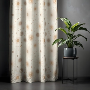 Op de afbeelding: Crèmekleurig gordijn met een herhalend patroon van zonnen, manen, sterren en kometen in tinten bruin en beige. Het gordijn hangt voor een grijze muur, met een plant in een pot op een kleine tafel rechts.
