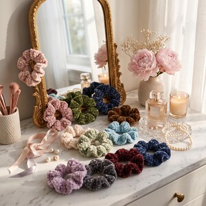May include: A collection of velvet hair scrunchies in various colors including pink, green, blue, brown, and purple, arranged on a white marble surface alongside a perfume bottle, a string of pearls, and a gold-framed mirror.