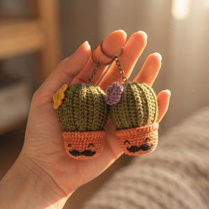 May include: Two crocheted cactus keychains featuring olive green bodies in peach-colored pots with embroidered black mustaches and smiling faces. One cactus has a small yellow flower, and the other has a small purple flower, both attached to metal rings.