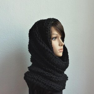 Könnte beinhalten: Ein schwarzer Strick-Hoodie und -Schal-Kombination. Das dicke, grobe Gestrick erzeugt ein warmes, strukturiertes Aussehen. Der Schal umschließt den Hals, während die Kapuze das Gesicht einrahmt. Dieses Winteraccessoire ist für Wärme und Stil konzipiert.