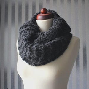 Könnte beinhalten: Ein dunkelgrauer, grob gestrickter Loop-Schal, der auf einer Schaufensterpuppe präsentiert wird. Der Schal besteht aus einem dicken, strukturierten Garn, das ein warmes und gemütliches Aussehen erzeugt. Der Schal ist um den Hals der Schaufensterpuppe gewickelt und zeigt sein kreisförmiges Design. Der Hintergrund hat vertikale Streifen.