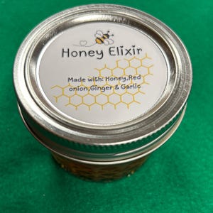 May include: A jar of Honey Elixir with a silver lid. The label features a cartoon bee and the words "Honey Elixir" above the ingredients: honey, red onion, ginger, and garlic. The jar is filled with a golden liquid.