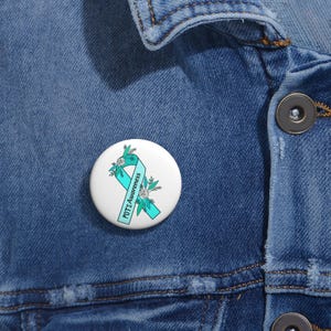 May include: A white button pin with a teal ribbon and floral accents. The text "POTS Awareness" is printed on the ribbon. The pin is set against a blue denim background.