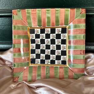May include: Square decorative plate with a checkerboard center featuring floral designs. The border has alternating stripes of orange and green, with gold trim. The plate is set on a pink satin surface.
