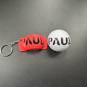 Peut inclure: Un porte-clés rouge avec le nom "PAUL" imprimé en noir, à côté d'une balle de golf blanche avec le nom "PAUL" en noir. Le porte-clés est rouge et possède un anneau argenté.
