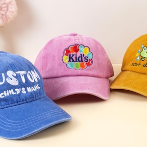 Op de afbeelding: Drie kleurrijke baseballpetten. Een blauwe pet met "CUSTOM CHILD'S NAME" in wit, een roze pet met "Kid's" en ballonnen, en een gele pet met een kikker en "your design".