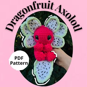 Puede incluir: Un ajolote de crochet de fruta del dragón en tonos rosa, verde y blanco. El ajolote tiene un cuerpo rosa brillante y está rodeado de rodajas de fruta del dragón verdes y blancas. También se ve el texto "Dragonfruit Axolotl" y "PDF Pattern".