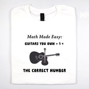 May include: White t-shirt with a black graphic of two guitars and the text "Math Made Easy: GUITARS YOU OWN + 1 = THE CORRECT NUMBER". The word "WHITE" is printed in the upper right corner.