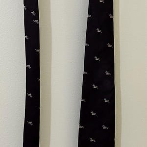 Puede incluir: Una corbata negra con un patrón repetido de pequeñas figuras de animales de color claro. La corbata cuelga de una percha de madera contra una pared blanca. Un accesorio clásico.