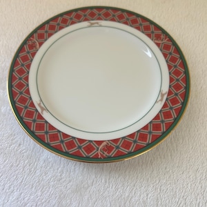 May include: A white ceramic plate with a red and green geometric border design. The border features a diamond pattern and small reindeer figures. The plate has a gold rim.