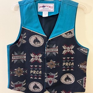Op de afbeelding: Een marineblauwe vest met een turquoise kraag en bies. De vest heeft een knoopsluiting en is versierd met rodeo-thema logo's en geometrische patronen. De vest heeft een label met "PRORODEO JACKETS".
