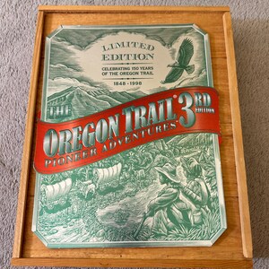 Peut inclure: Une impression encadrée commémorant le sentier de l'Oregon. L'impression présente des illustrations vertes et blanches de pionniers et de chariots. Le texte comprend "Édition Limitée" et "Célébrant 150 ans du sentier de l'Oregon 1848-1998". Le cadre est en bois clair.